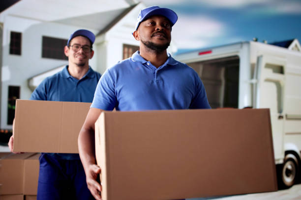 Professional team members in blue uniforms and caps carrying heavy cardboard packages forward from a moving van, demonstrating reliable services from movers and packers Airport Road.