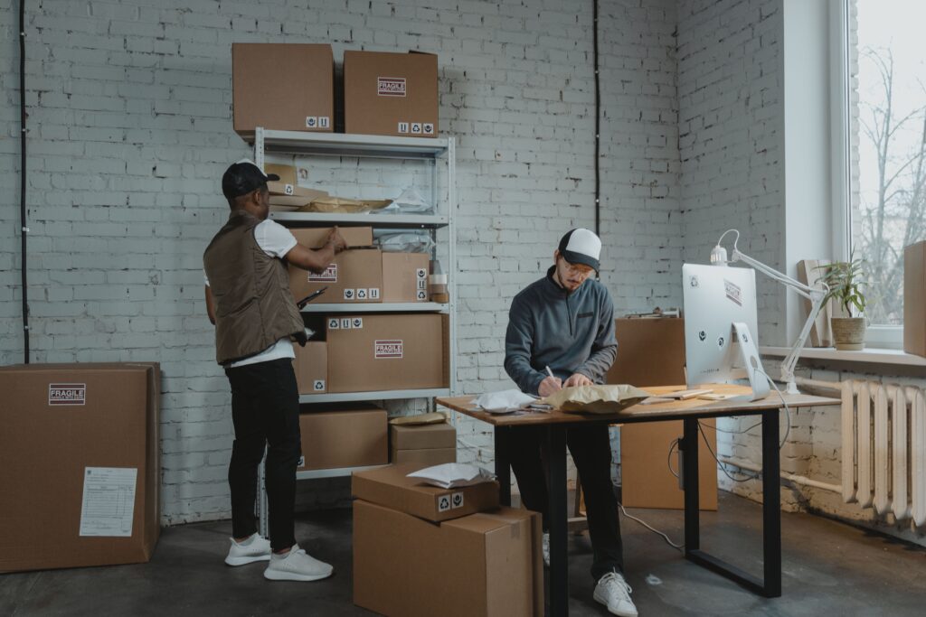 Two logistics workers organizing fragile cardboard boxes on shelving units and managing inventory at a desk, representing services by Abu Dhabi movers and packers.