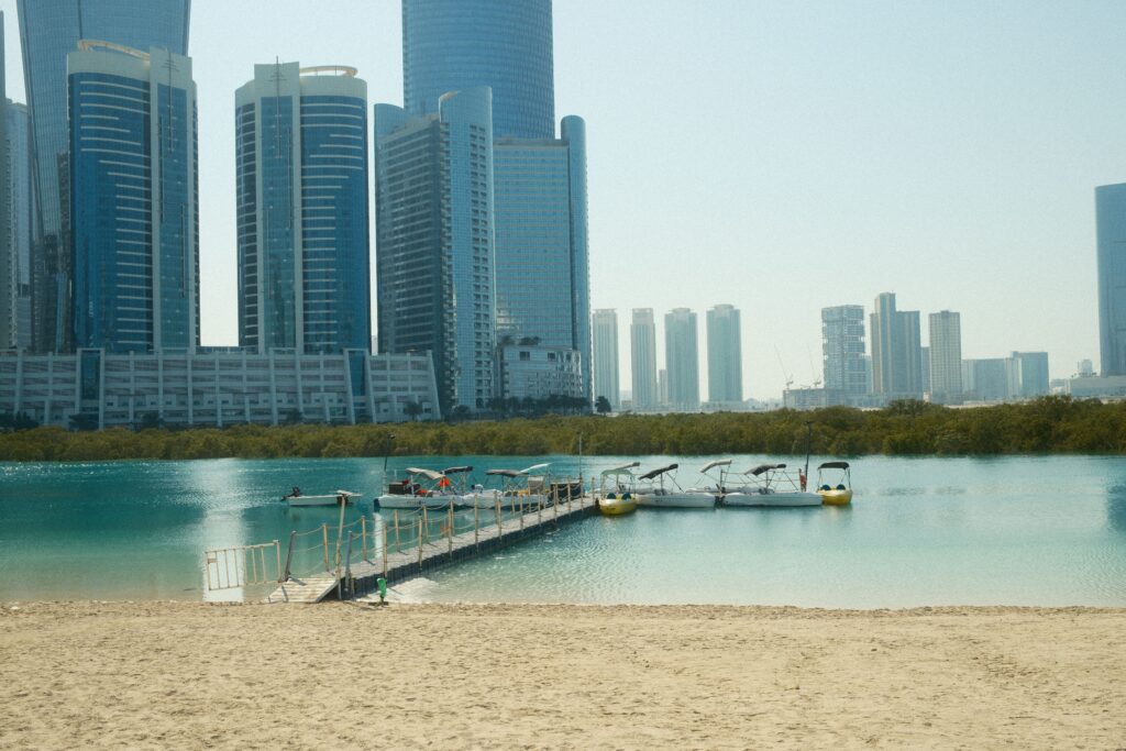 Panoramic view of modern waterfront skyscrapers, a sandy beach shoreline, and a wooden dock pier over clear water, highlighting service zones for movers and packers Al Raha Beach.