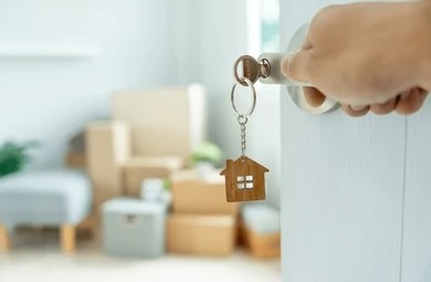 Hand unlocking a new villa door with a wooden house keychain against blurred moving boxes.