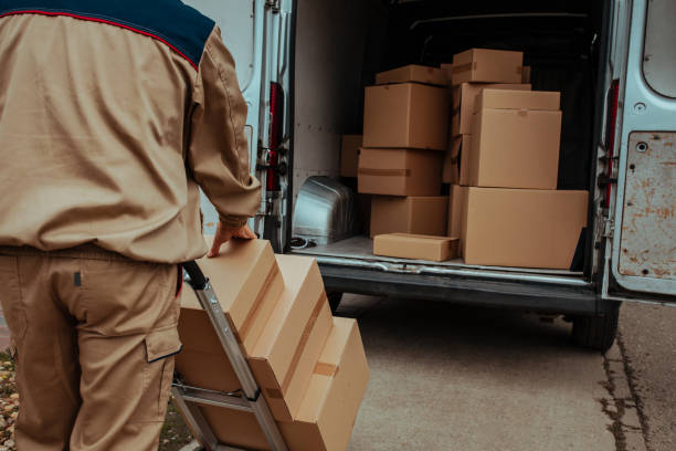 A relocation specialist pushing a hand truck stacked with cardboard boxes toward an open delivery van, illustrating services by movers and packers Al Raha Beach.