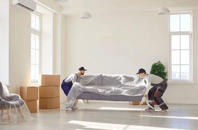 Professional movers carrying wrapped sofa in bright living room near cardboard boxes.