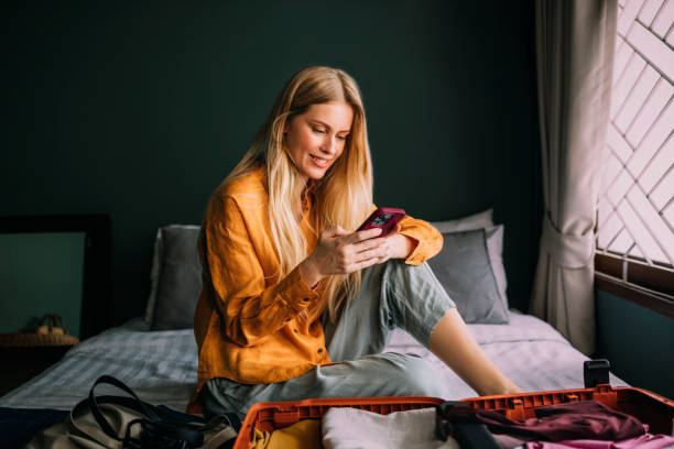 Young woman sitting on a made bed using her smartphone next to an open suitcase.