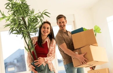 Smiling couple carrying moving boxes and a potted houseplant into their new home.