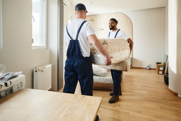 Two professional movers carrying a wrapped mattress inside a studio apartment.