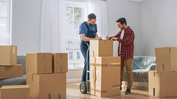Homeowner signing delivery paperwork on clipboard held by mover in living room filled with boxes.