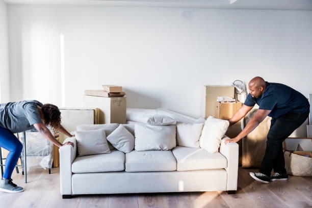 Two people moving a white fabric sofa into a bright apartment surrounded by packing boxes