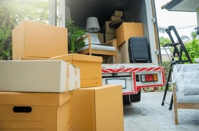 Stacked cardboard moving boxes in front of an open truck loaded with household furniture.